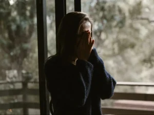 A woman standing in front of a window holding her face in her hands.