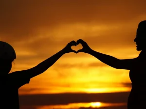 Two people holding their hands up to shape a heart against sunset.