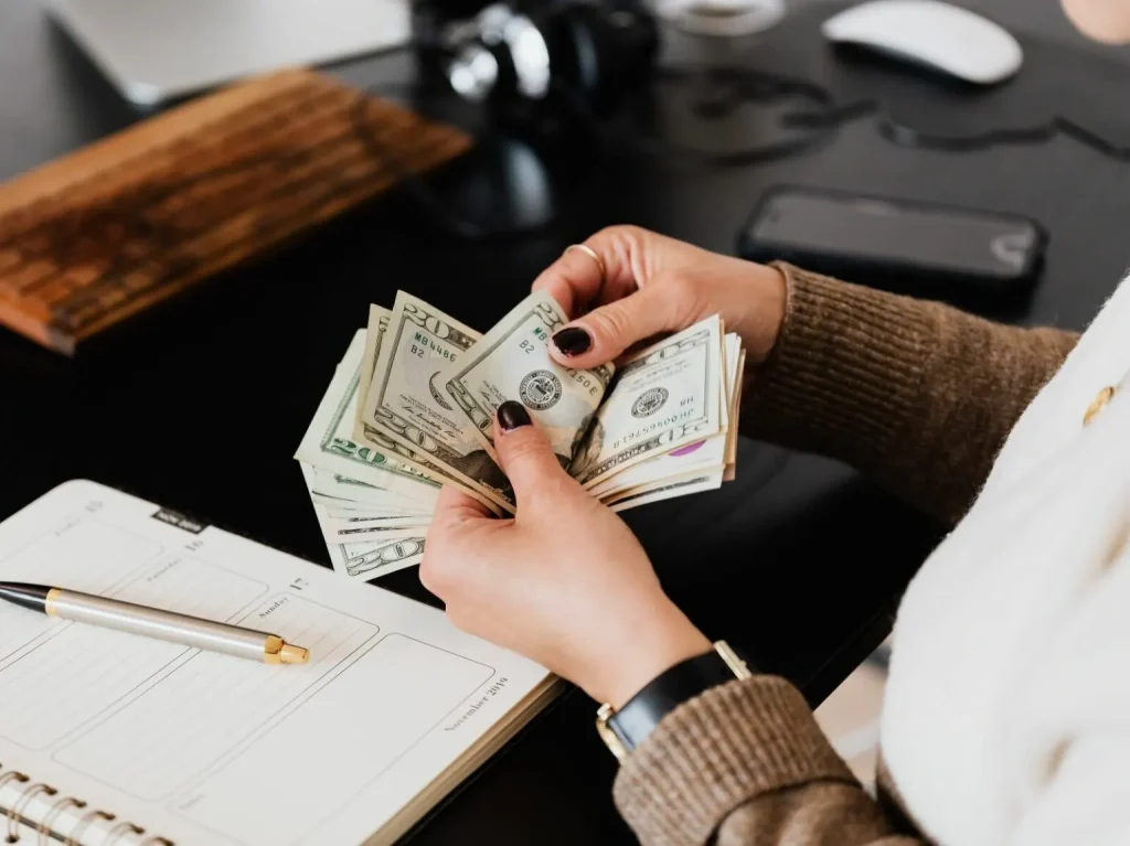 A woman counting money with a planner and cell phone on her desk.