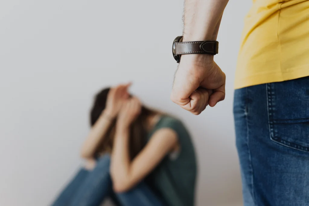 A woman sitting and shielding her face as a man angrily clinches his fist.