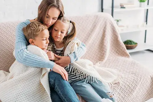 A mom holding her two kids on the couch with a blanket wrapped around them.