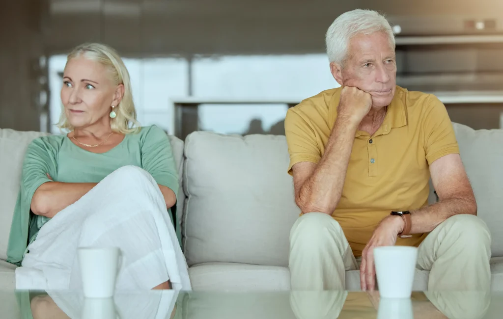 An older couple sitting on the couch looking upset.