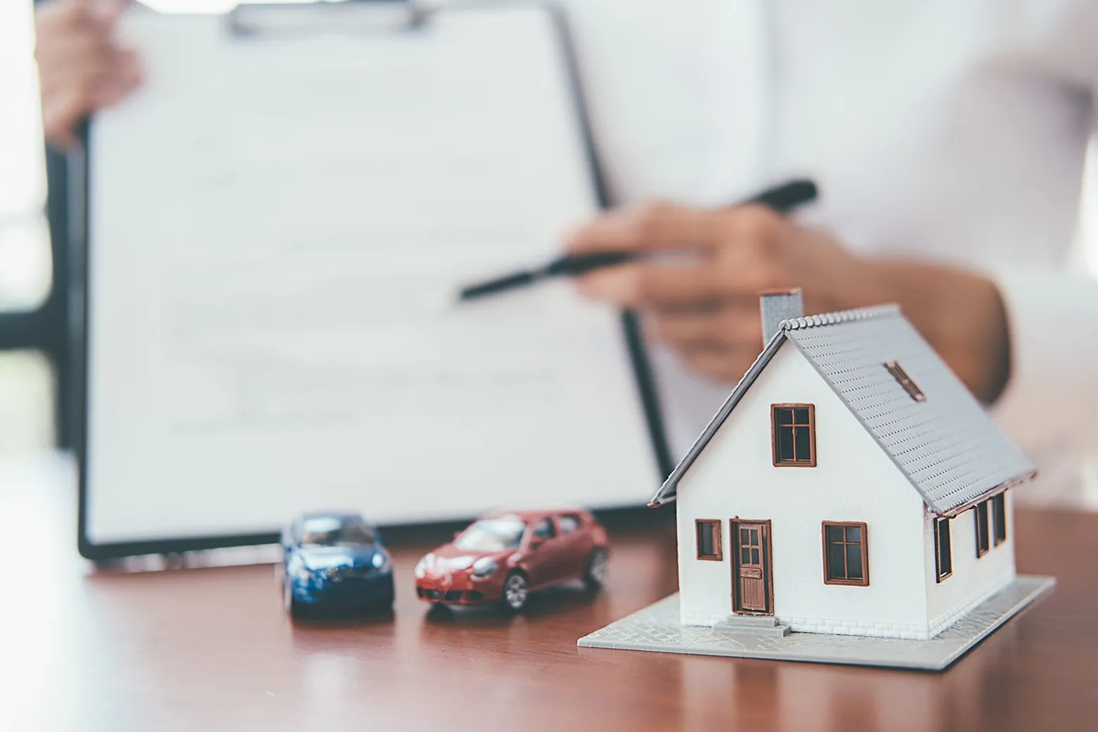 A house and cars on a desk with someone holding up paperwork in the background.