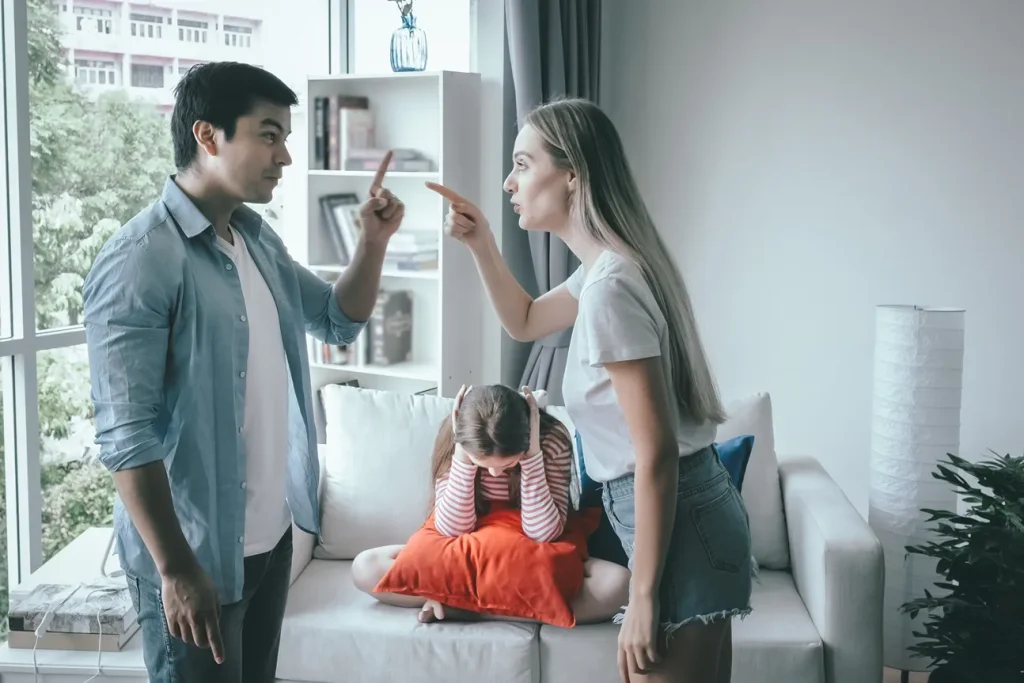 Parents arguing with each other with a child holding her ears in the background.