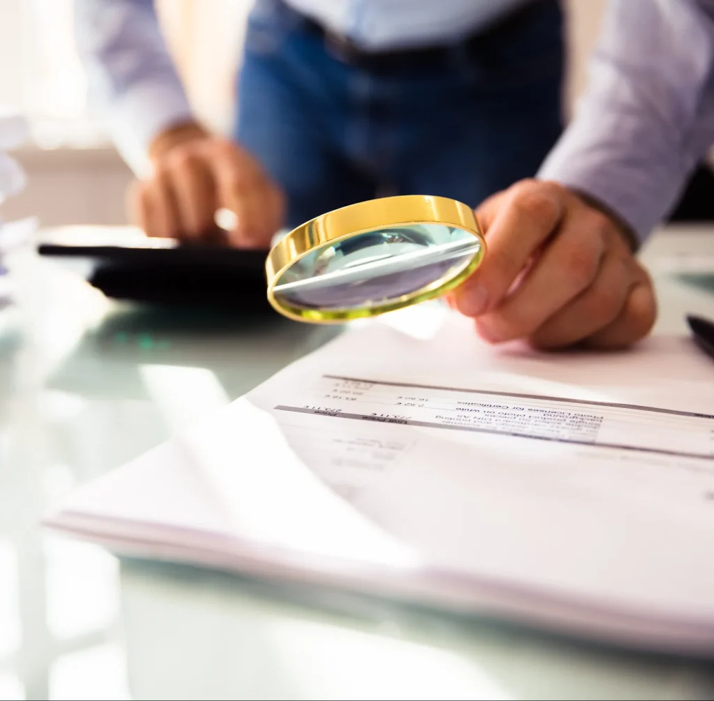 A man holding a magnifying glass up to accounting paperwork