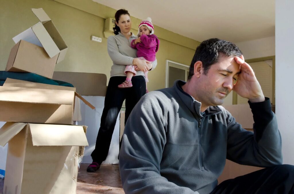 A mom holding her child, a stack of empty boxes, and a dad with his hand on his forehead.