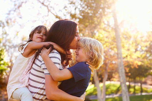 A mother with her daughter on her back and holding her son while kissing his head.