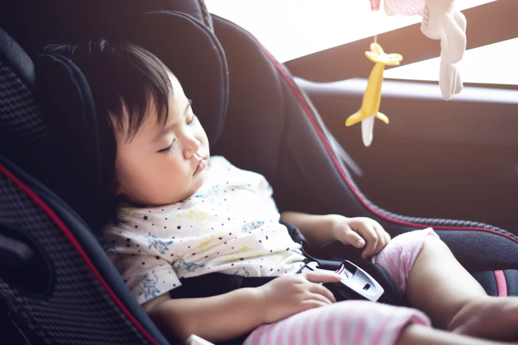 Young child sleeping peacefully in a car seat, wearing a patterned shirt and striped pants, with soft hanging toys above.