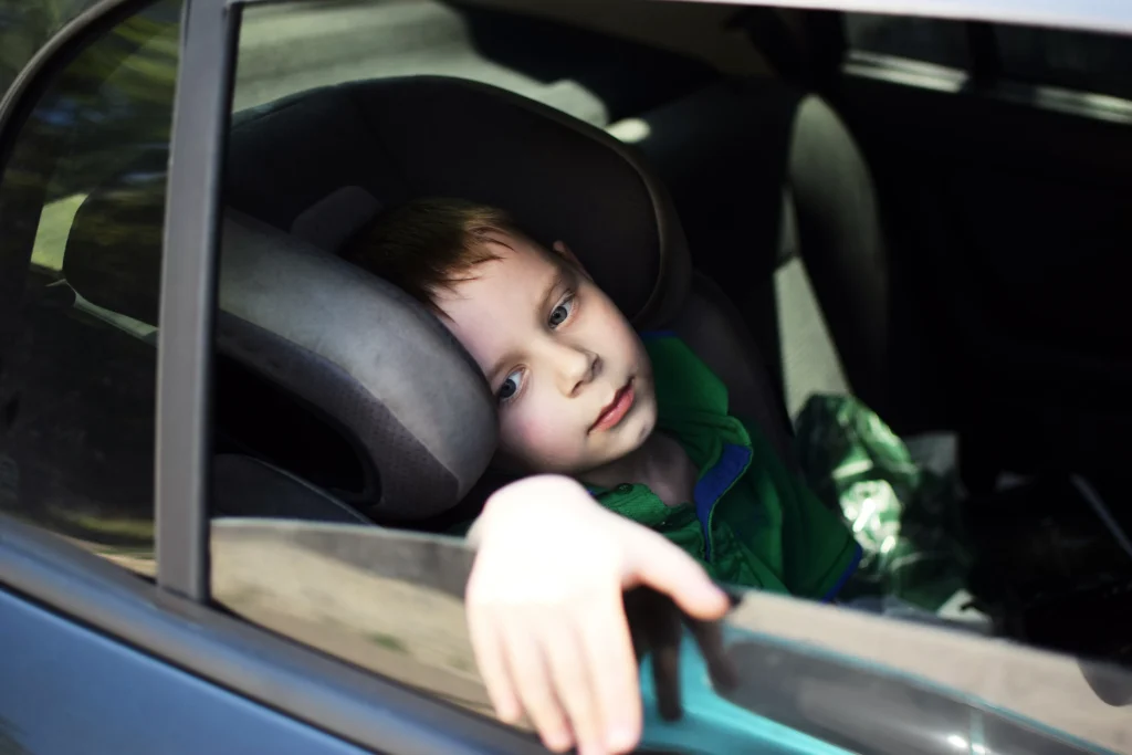 Young boy resting his head on a car seat, gazing out the window with a sad expression, wearing a green shirt and sitting in the back seat of a vehicle.